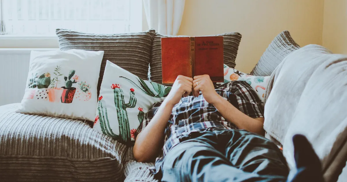 Relaxing at home, lying on a sofa with a book and cushions around.