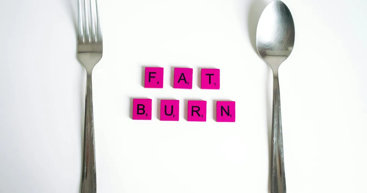 Conceptual art featuring the words 'Fat Burn' using Scrabble tiles between a fork and spoon on a white background.