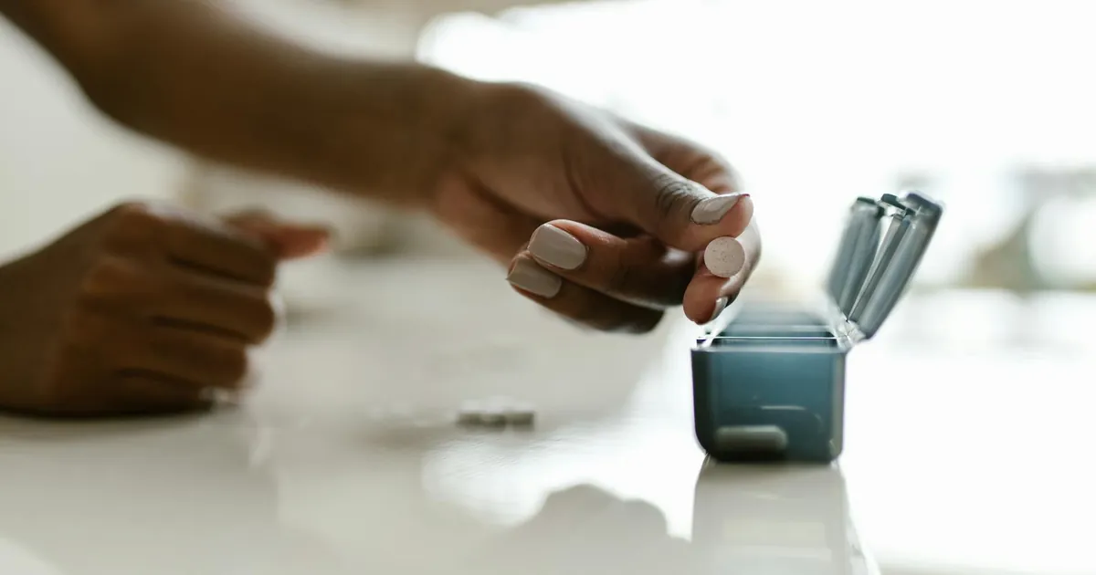 Hand placing pills in a weekly organizer, emphasizing medication management. Hand placing pills in a weekly organizer, emphasizing medication management.