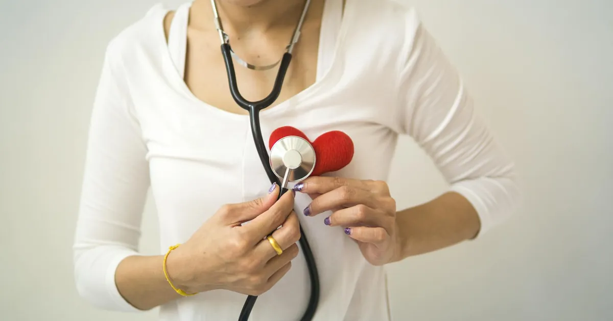 Unrecognizable female wearing white shirt while standing on white background with diaphragm of stethoscope on red handmade heart in room