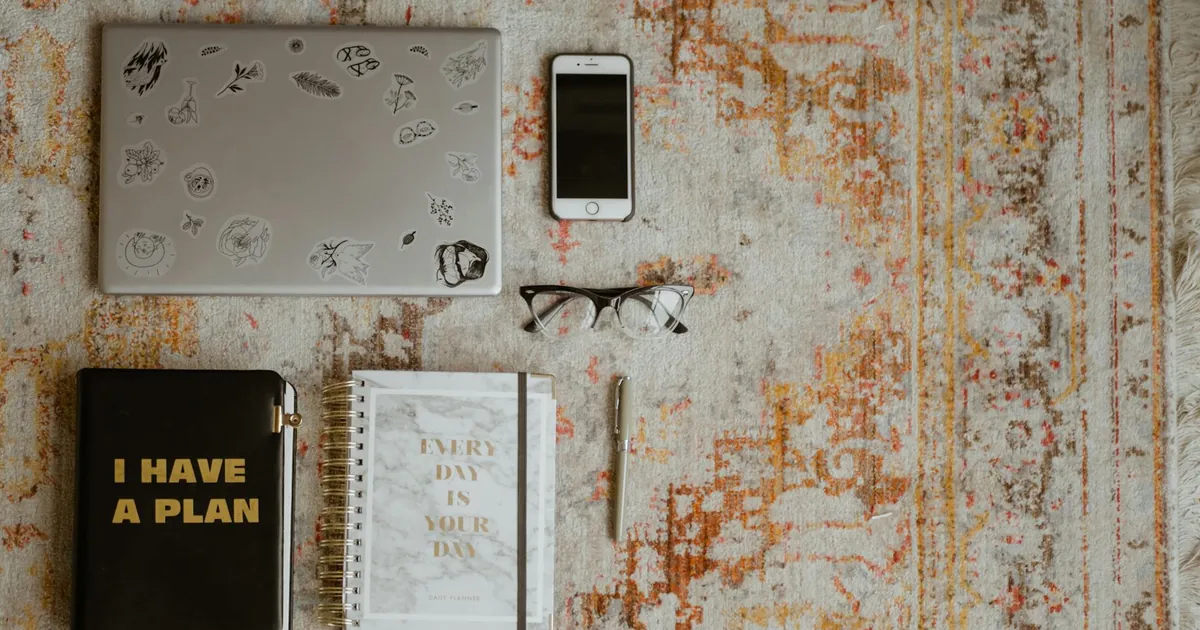 Flat lay of workspace items including laptop, phone, planners, and glasses on a patterned rug.