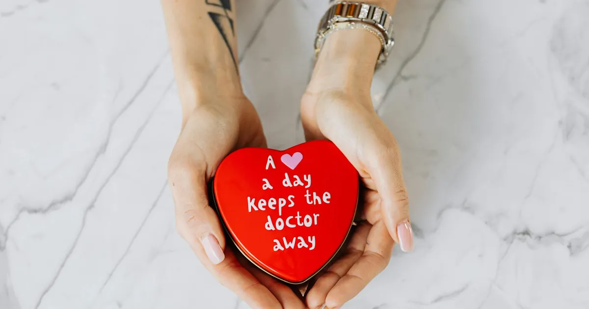 Hands holding a red heart-shaped tin with the message 'A heart a day keeps the doctor away' on a marble surface.
