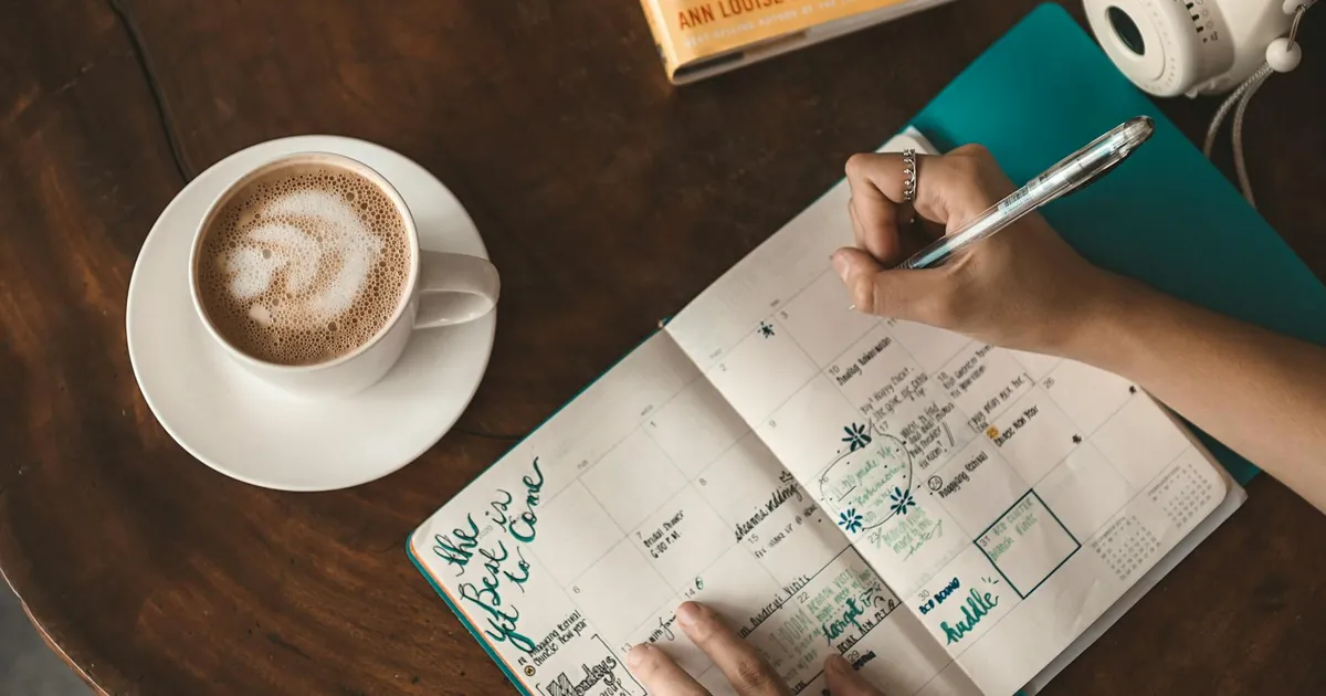 Woman writing in planner with cappuccino, book, and camera on wooden table. Woman writing in planner with cappuccino, book, and camera on wooden table.